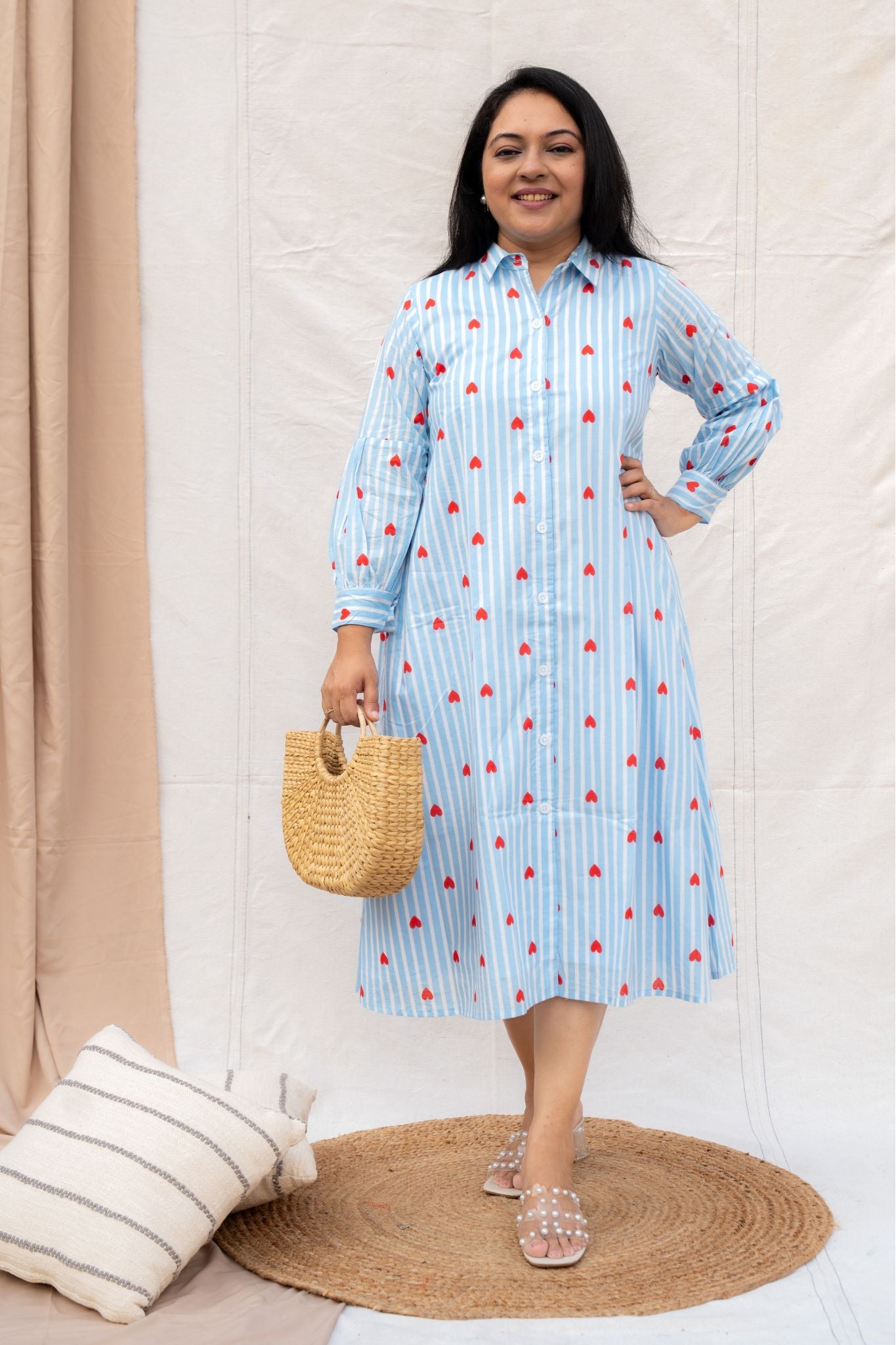 Woman wearing a blue dress with red patterns, holding a straw bag, standing on a beige rug.