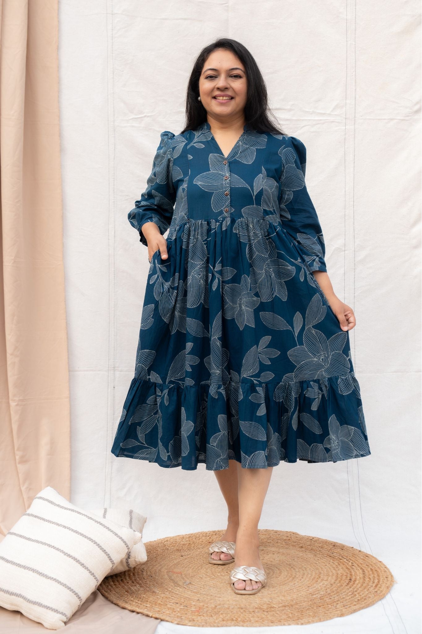 Woman wearing a blue floral dress standing in a room with beige curtains and a white wall.