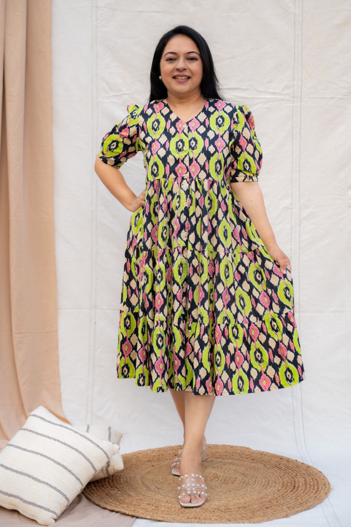 Woman wearing a colorful patterned dress standing against a white wall with a beige curtain.