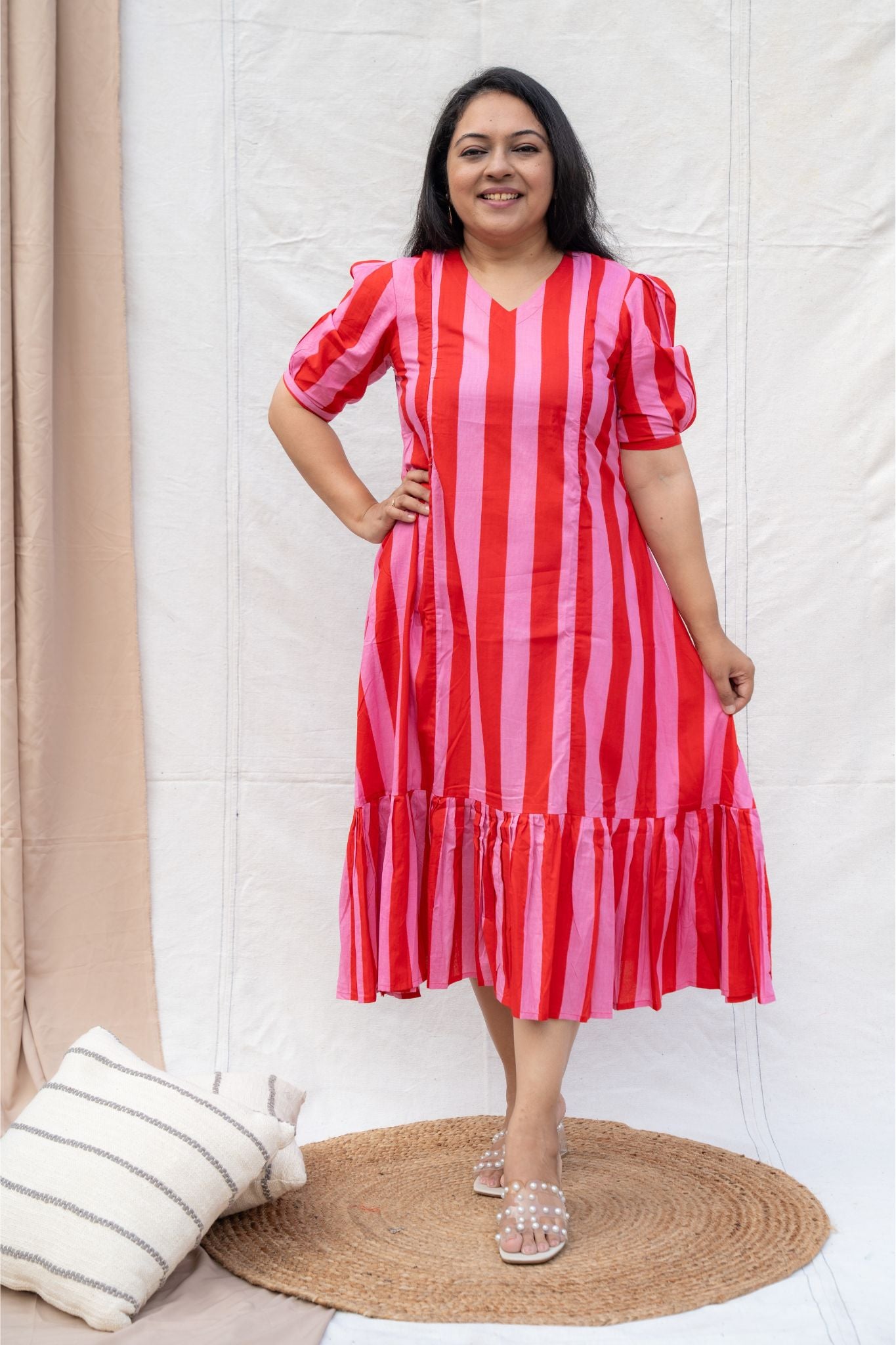 Woman wearing a red and pink striped dress standing in a room with a white wall and beige curtain.