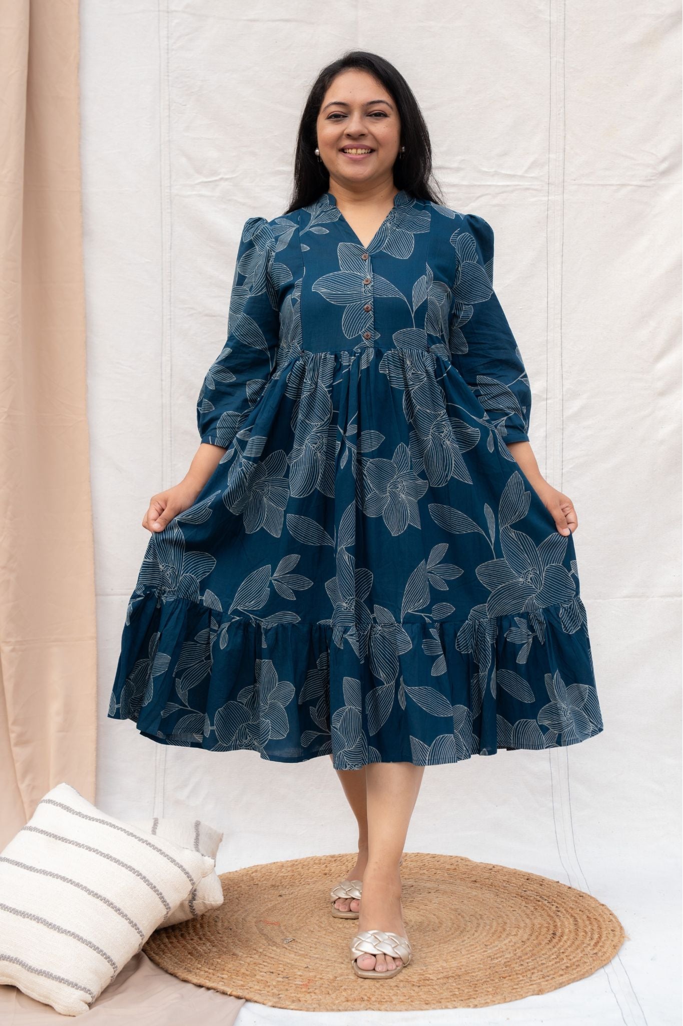 Woman wearing a blue floral dress standing in a room with a white wall and beige curtain.